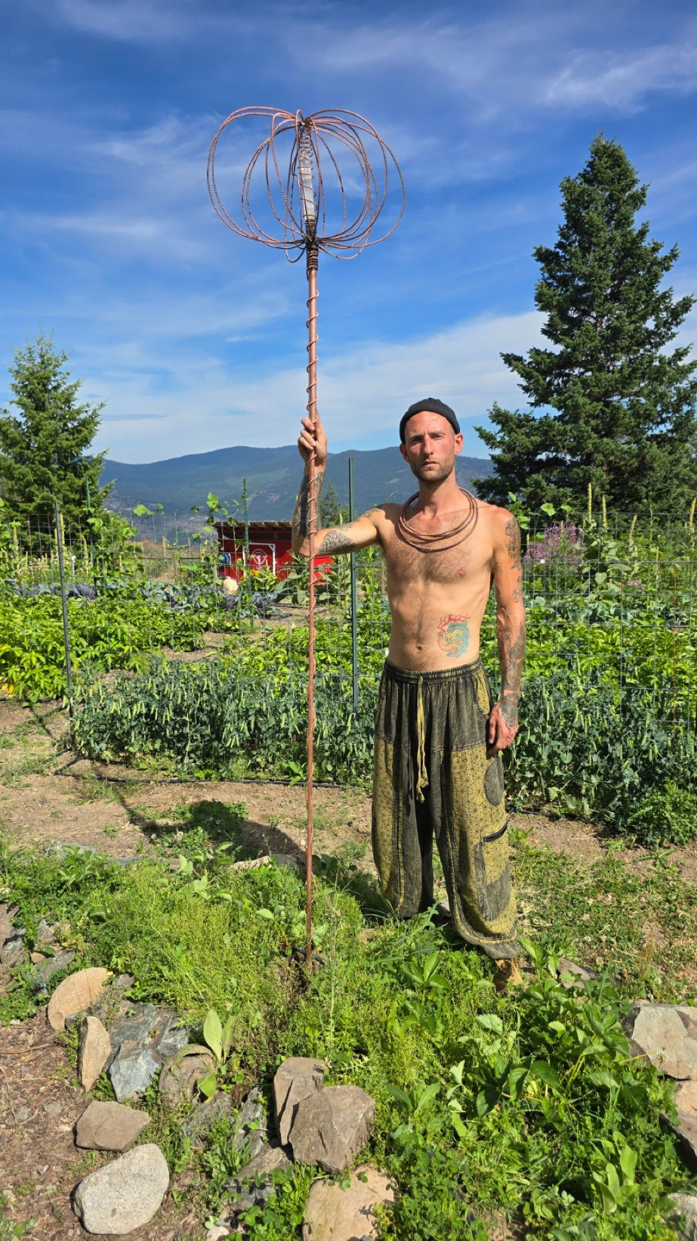 Steven standing in the garden holding the Electroculture Harmonizer Antenna System, a hand-forged copper crown set with a quartz crystal on its twisted-copper rod, lit against a dark ground